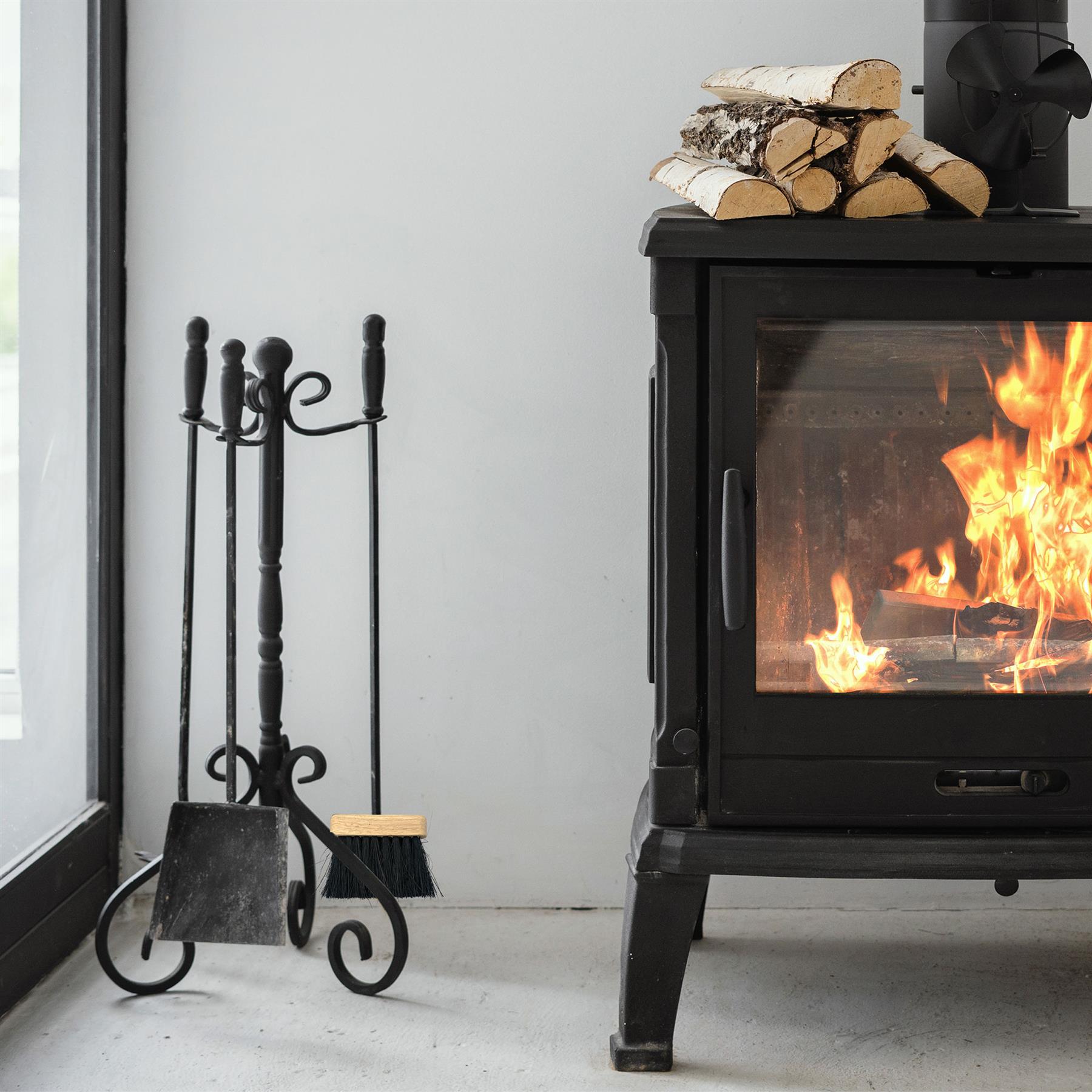 Black wrought iron fireplace tool set with a poker, shovel, and brush, displayed next to a black wood-burning stove with a visible fire and stacked birch logs.