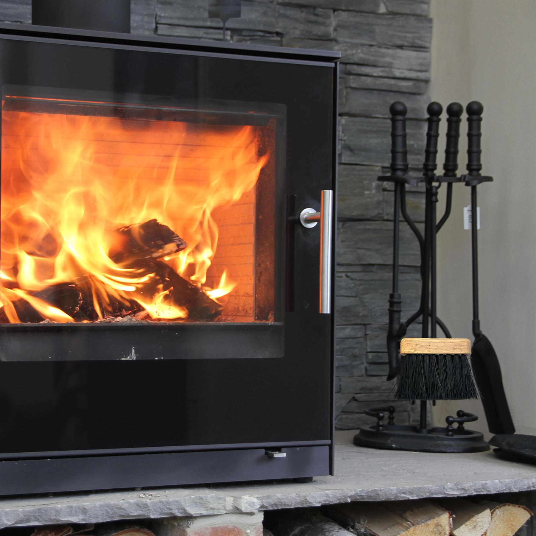 A black wood-burning fireplace with bright orange flames burning inside, set on a stone hearth next to a black metal fireplace tool set against a gray stone wall.