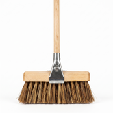A traditional push broom with a light wood handle, a wooden head, and natural brown bass and cane bristles, featuring a silver metal connector.