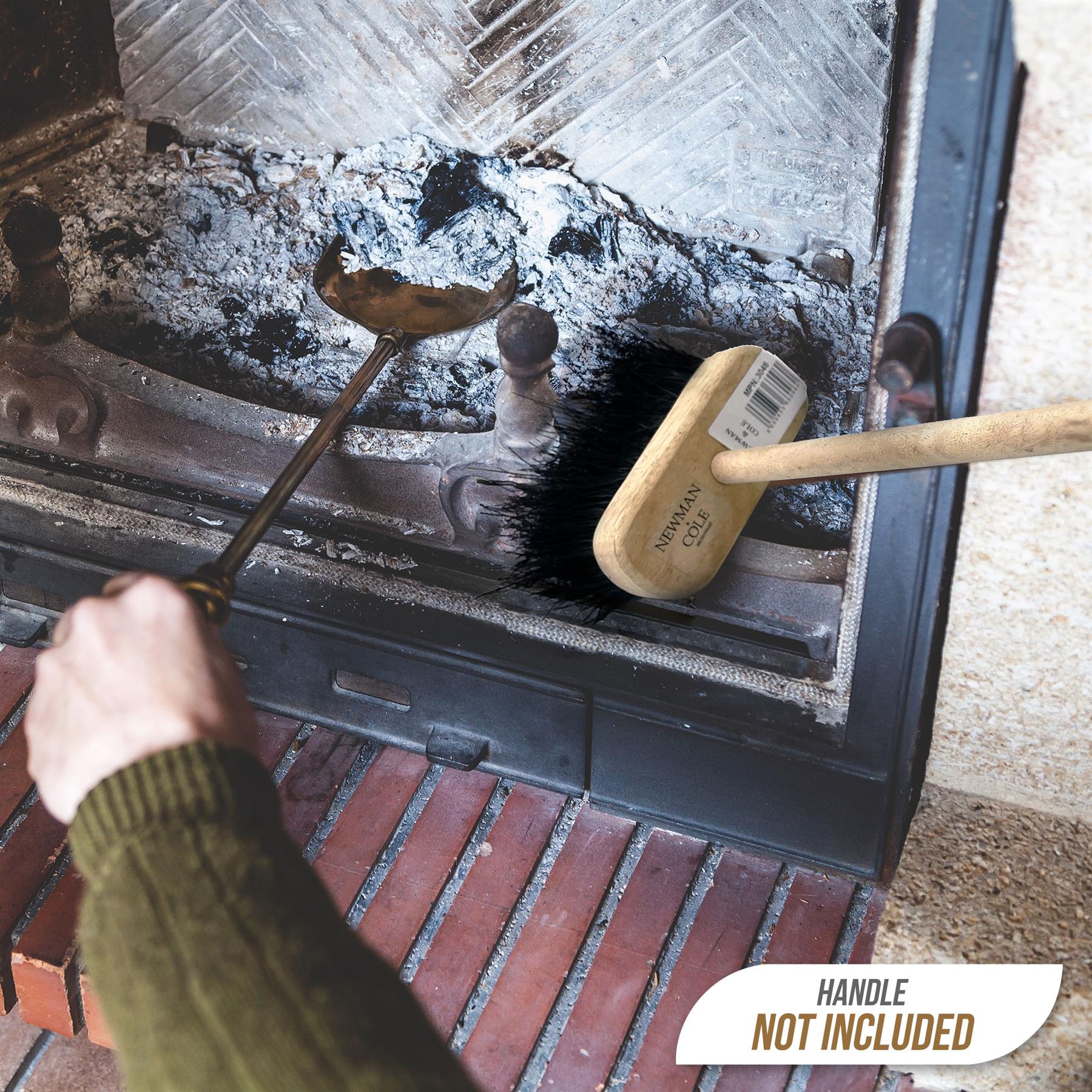 A close-up of a fireplace or grill interior with a person's hand using a metal tool, alongside a brush with a light wood handle. A white badge in the corner reads 'HANDLE NOT INCLUDED'.