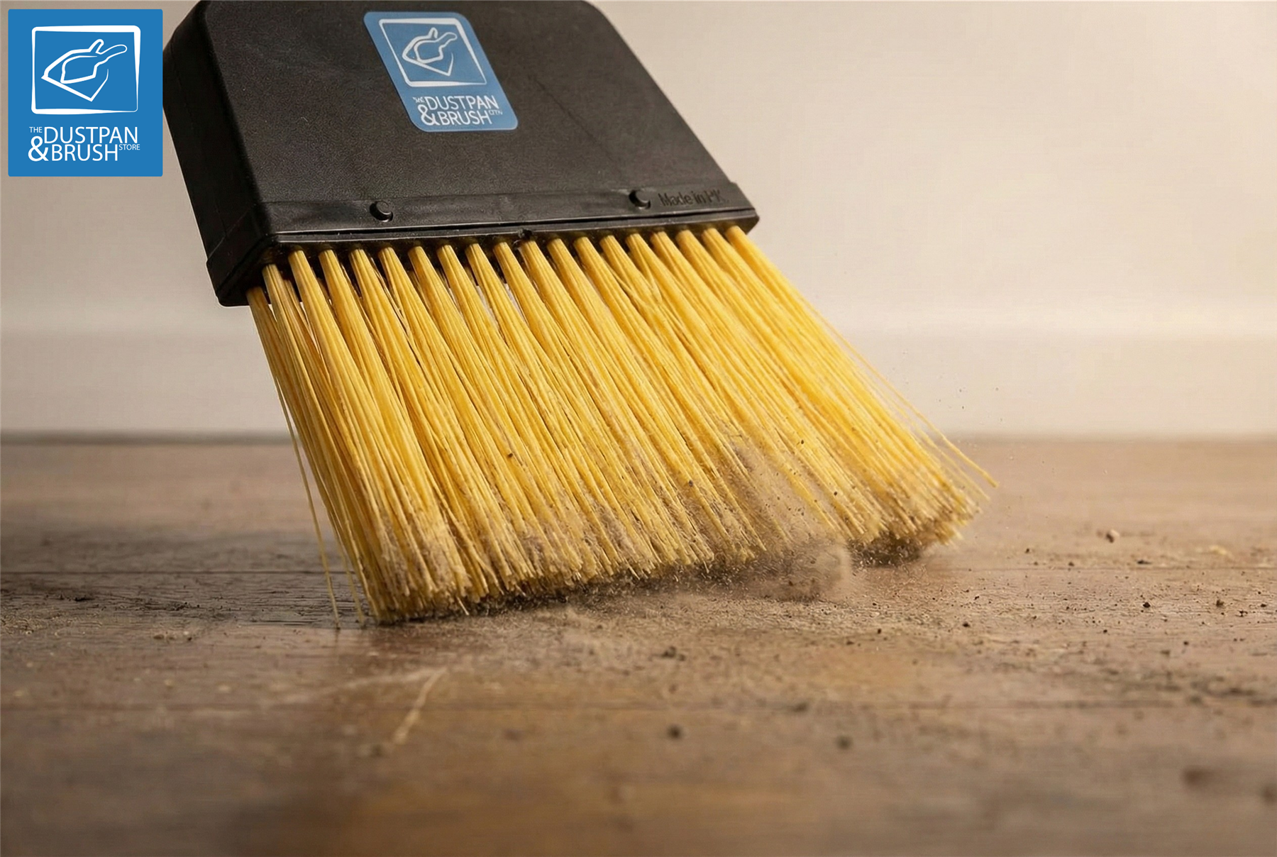 A close-up of a dustpan brush with a black handle and golden-yellow bristles, shown sweeping dust and debris across a wooden floor. 