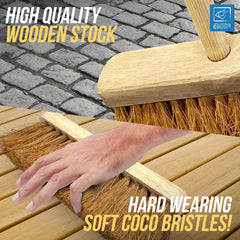 A close-up promotional shot of a wooden broom with soft coco bristles being swept across a light wood surface, with a human hand resting on the bristles for scale. Text overlays read 'HIGH QUALITY WOODEN STOCK' and 'HARD WEARING SOFT COCO BRISTLES!' 